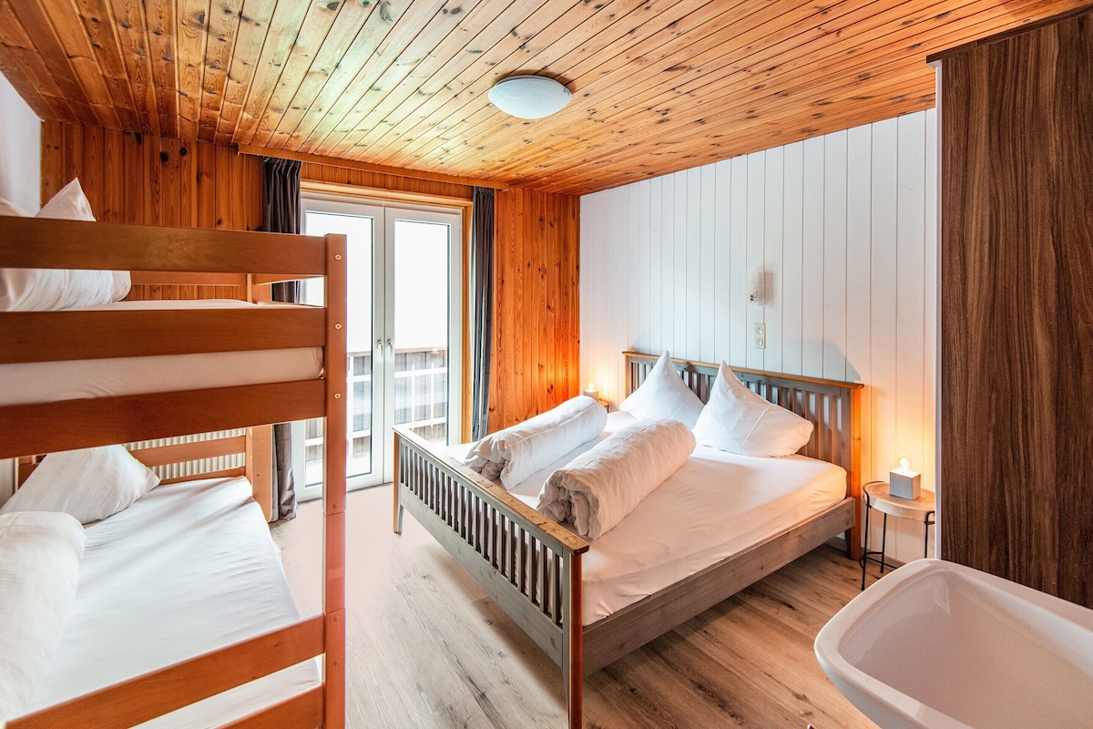 A wooden cabin room with bunk beds, a single bed, a bedside table, a nightstand, and a window.