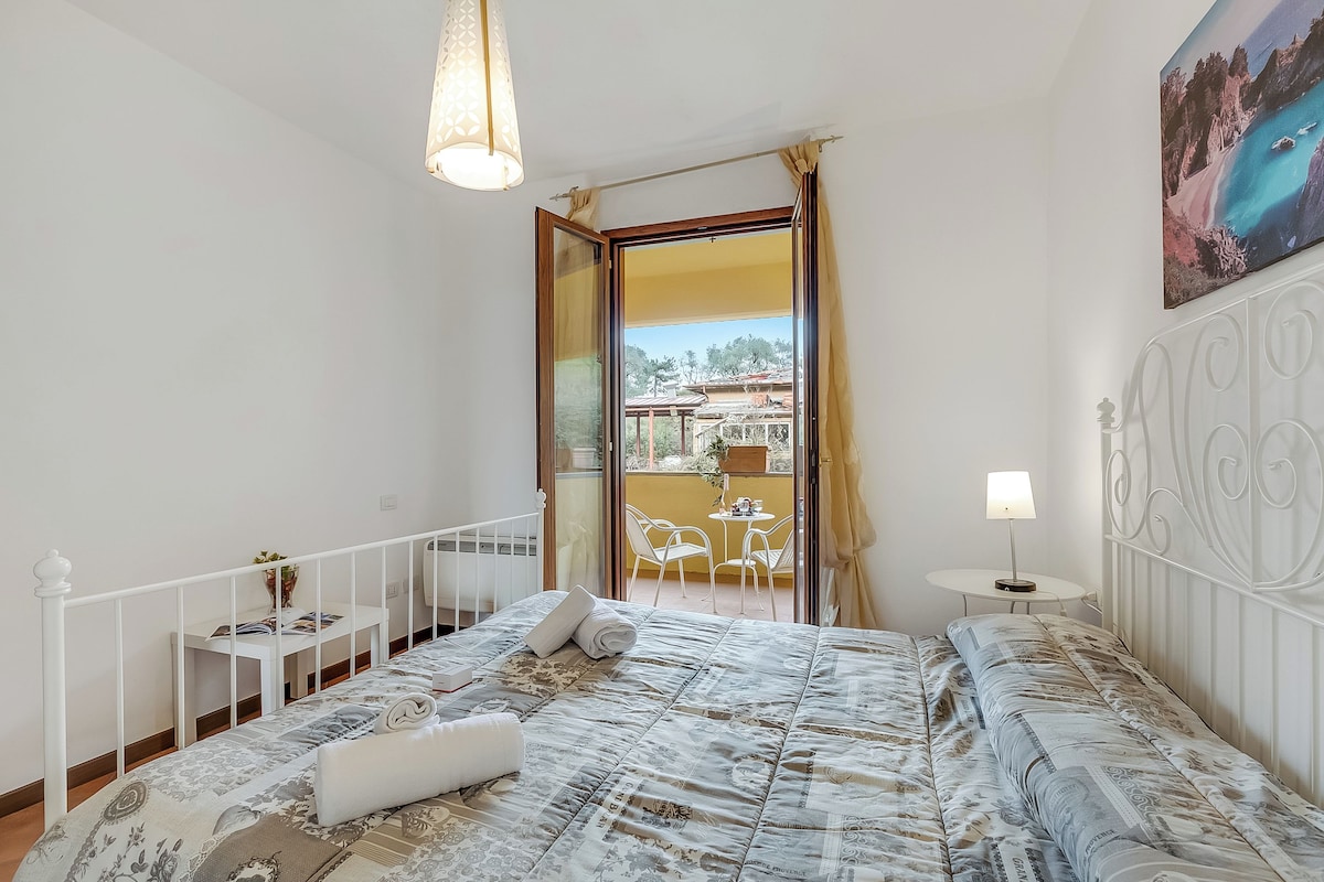 A bedroom with a bed, a bedside table, a lamp, and a view of a balcony with outdoor furniture.