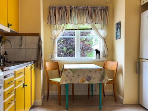 Una cocina con armarios amarillos, una ventana con cortinas, una mesa con mantel floral y un perro negro en el alféizar.