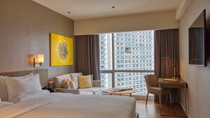 A hotel room with a large bed, a desk, a chair, a sofa, a window with a city view, and a wall-mounted artwork.