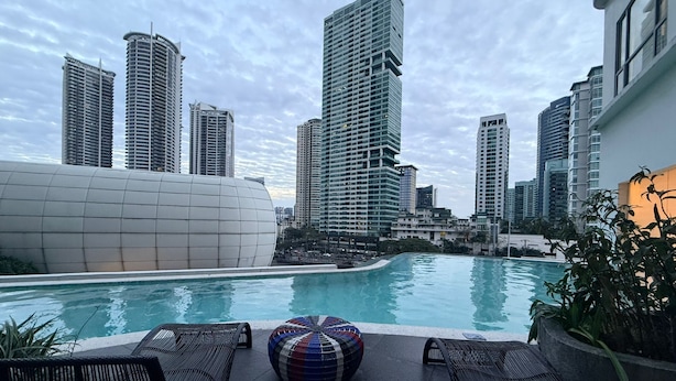 A rooftop pool with lounge chairs and a colorful ottoman, surrounded by high-rise buildings.
