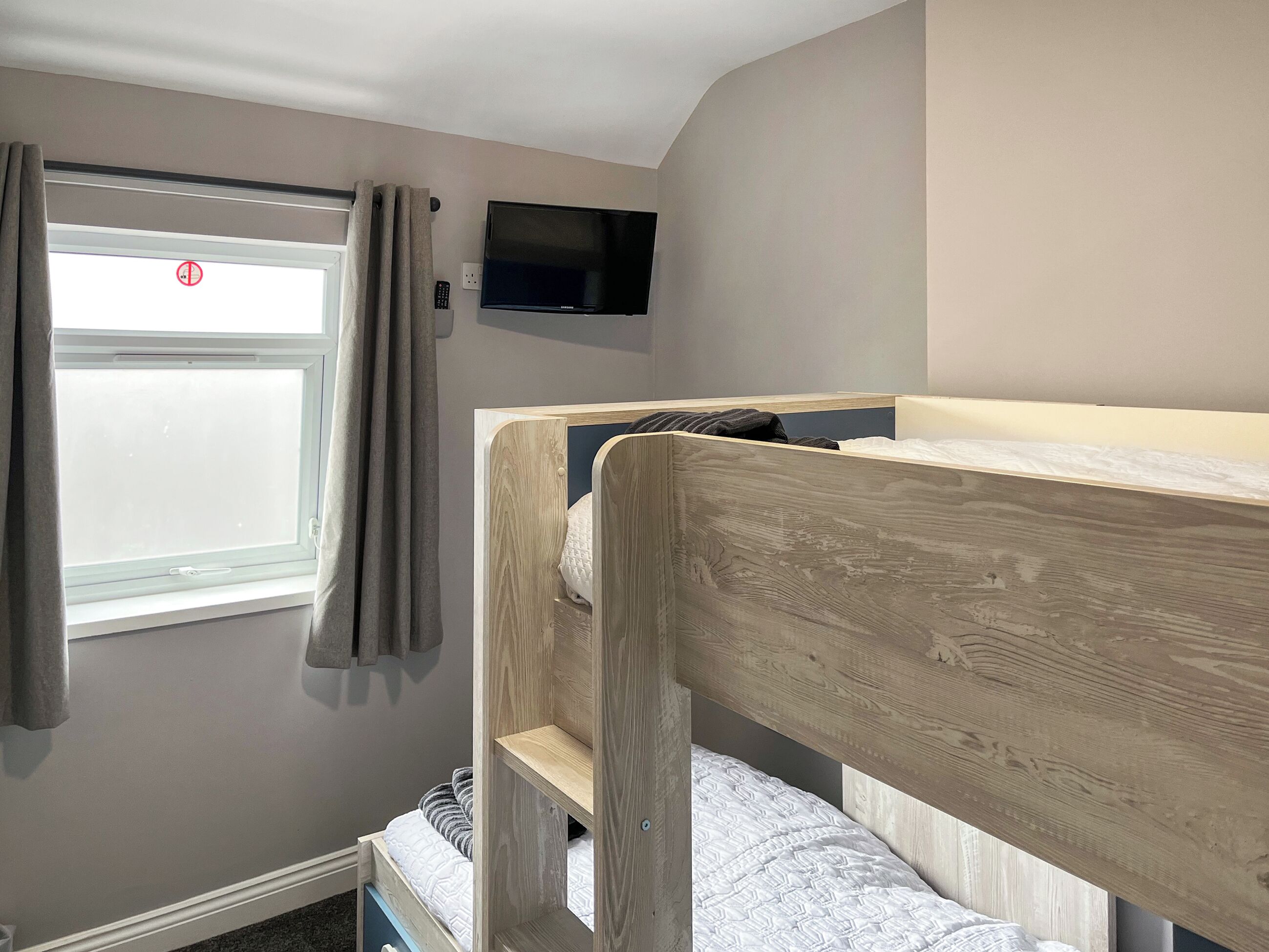 A bunk bed with a television above it, a window with curtains, and a wall-mounted light switch.