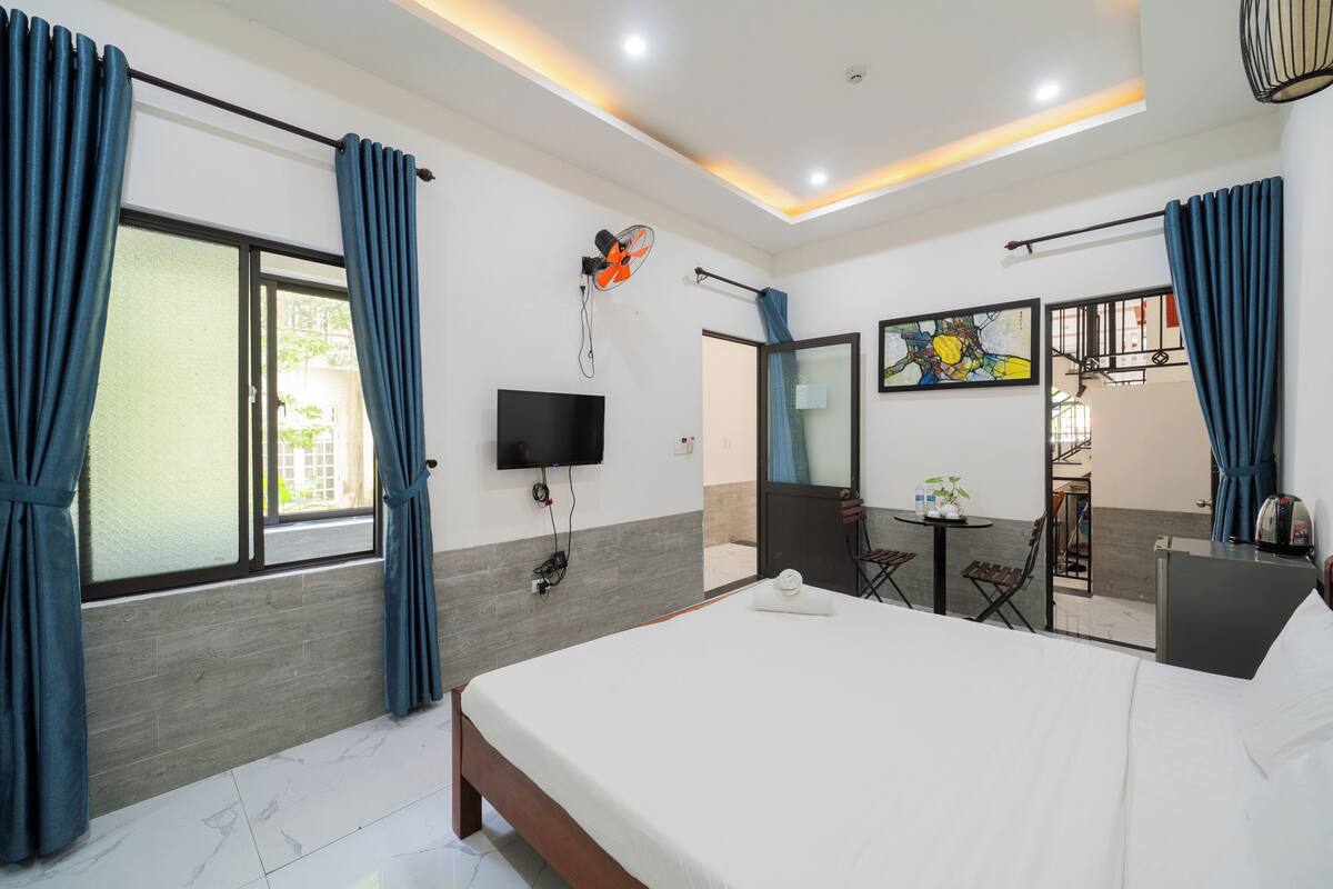 A hotel room with a bed, a TV, a fan, a small table with a plant, and a view of the outside.