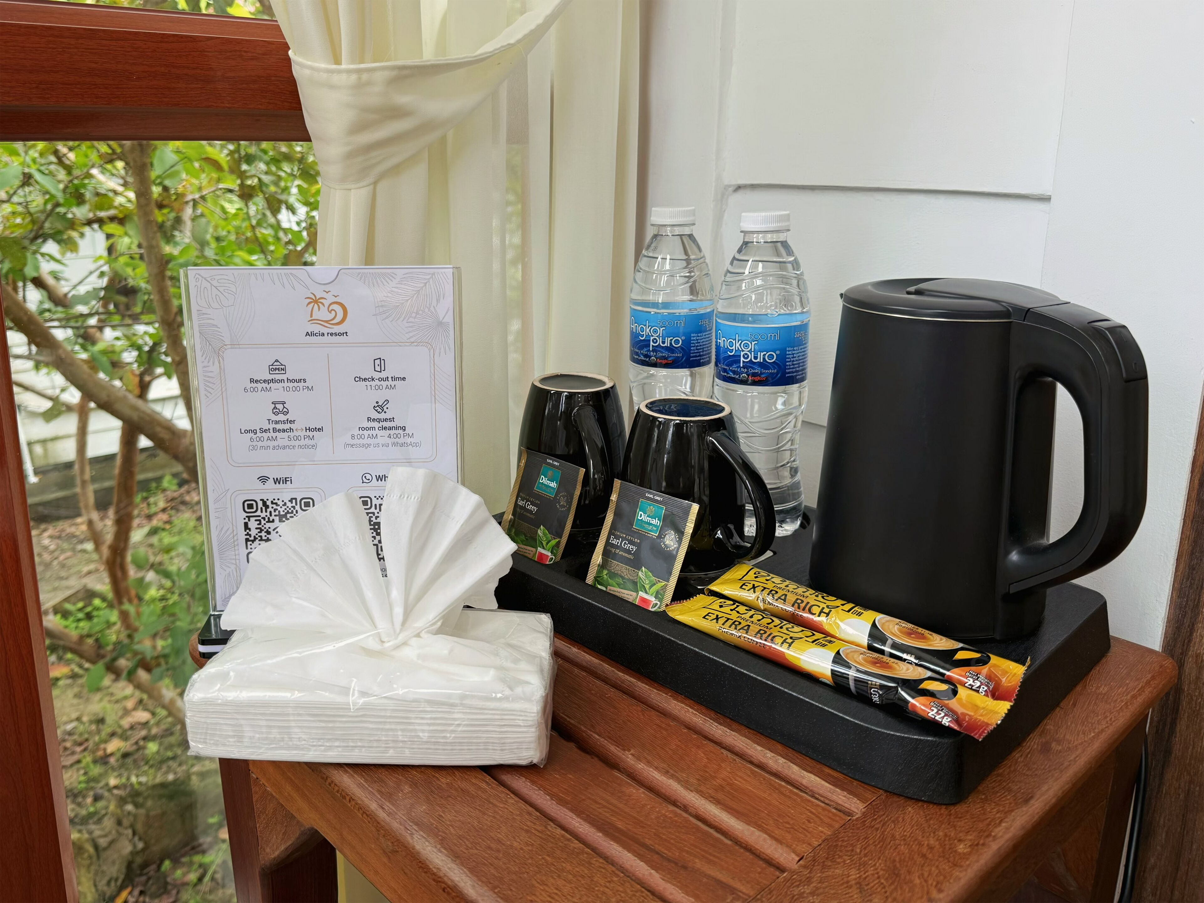 A hotel room with a wooden side table, a tray with coffee and tea packets, a black coffee pot, two water bottles, and a room service menu.