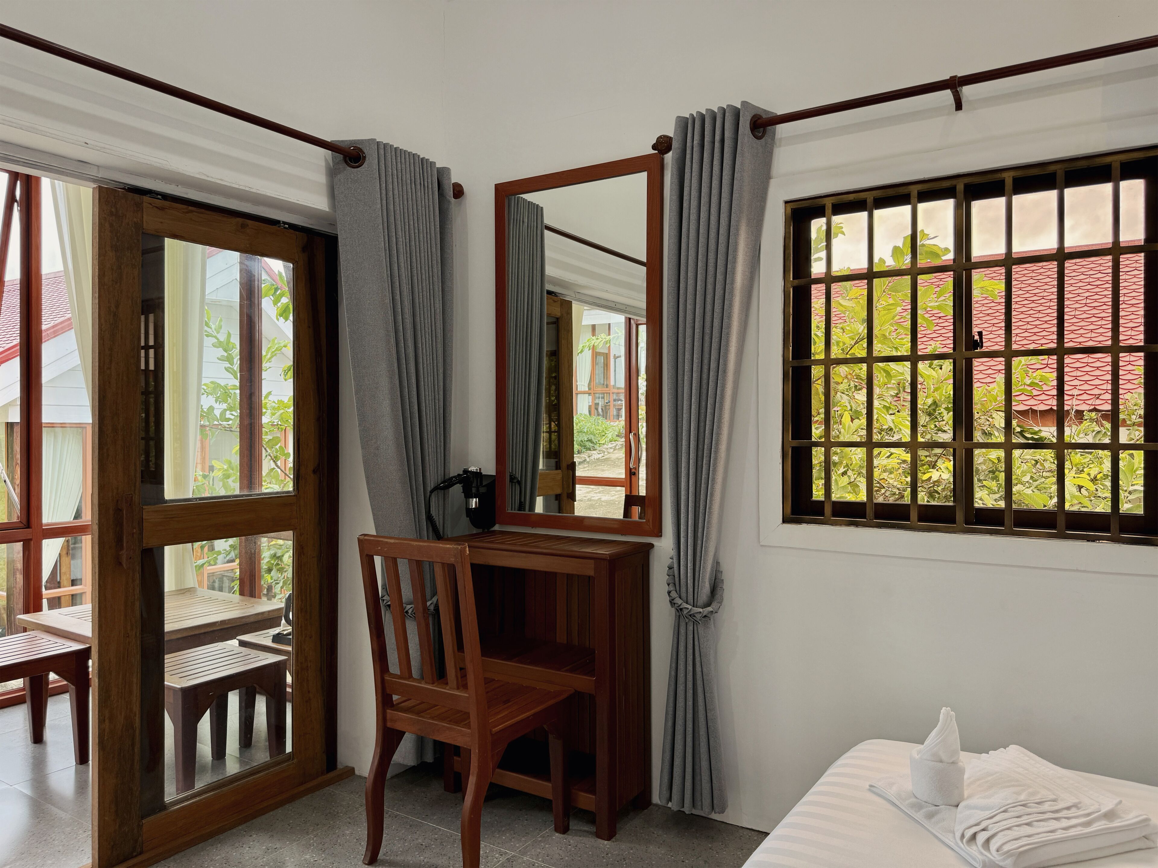 A room with a wooden desk, a chair, a bed, and a window with a view of greenery and a red-roofed building.