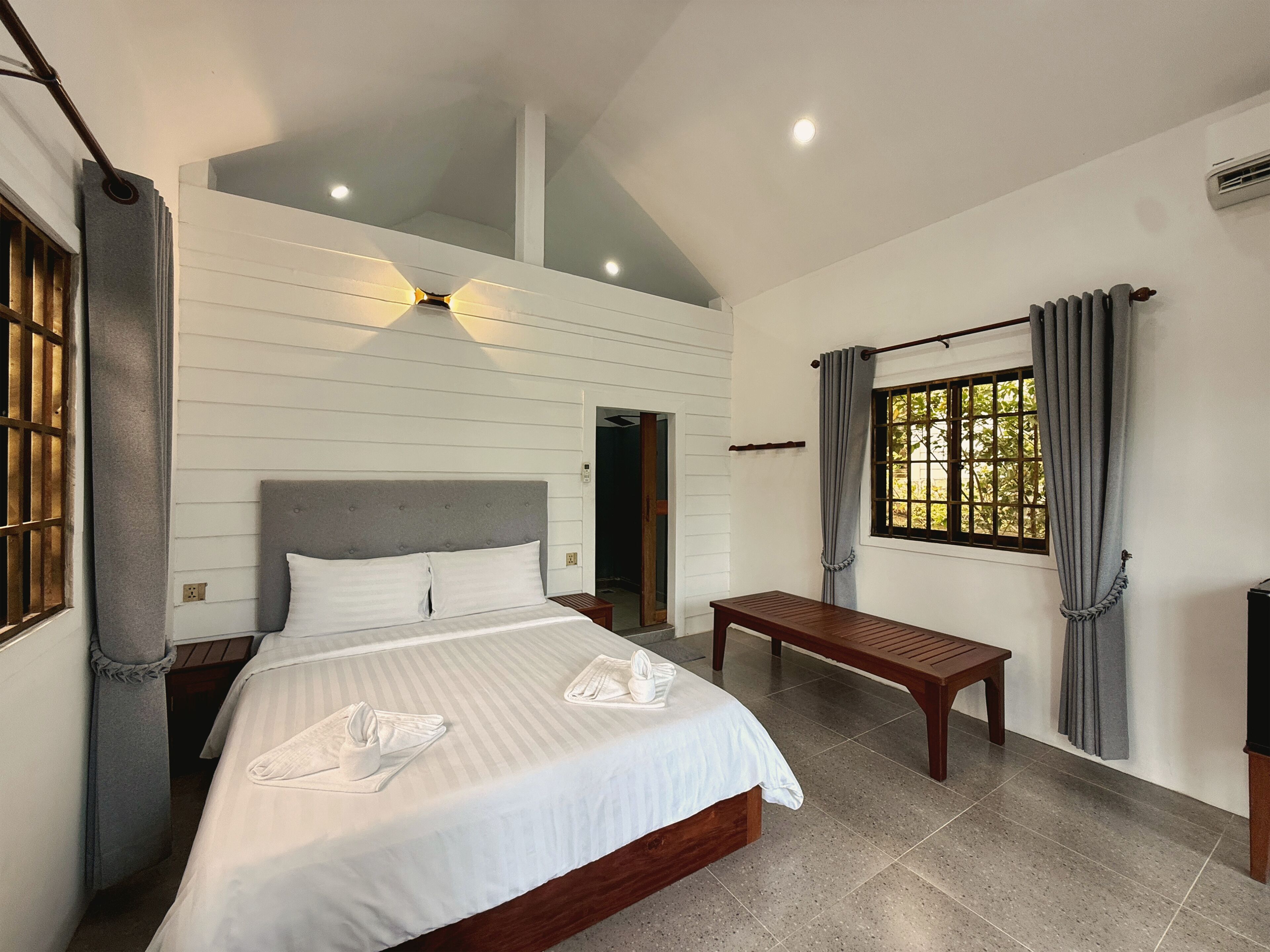 Un dormitorio con cama, una banca, una ventana con cortinas y una lámpara de techo.