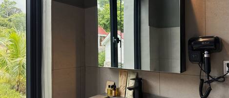 A bathroom with a large window, a sink, a mirror, and a hairdryer mounted on the wall.