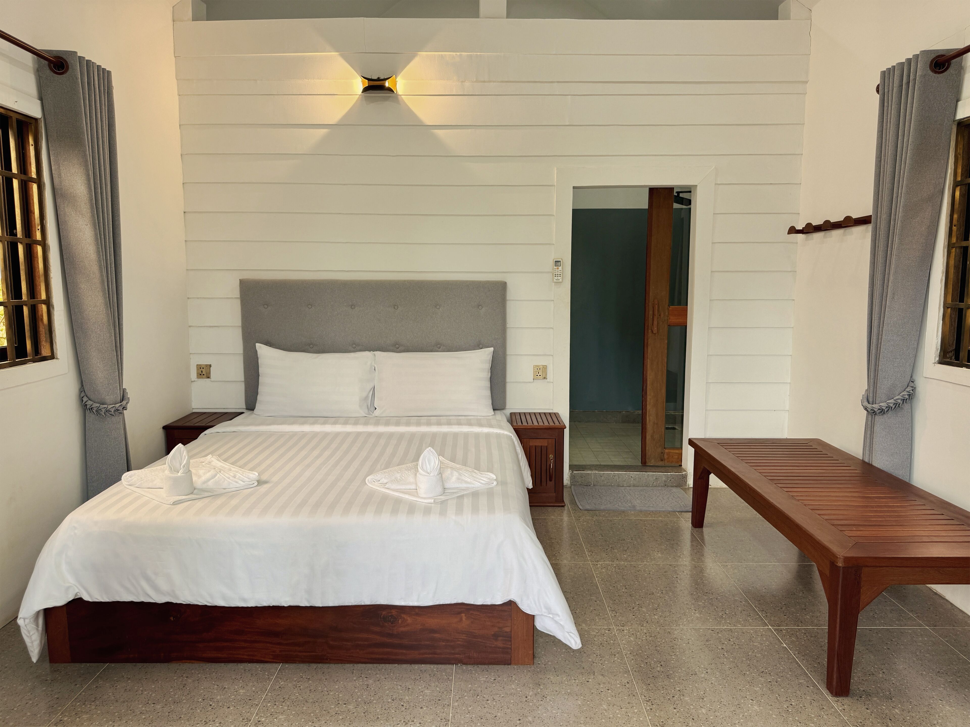 A bedroom with a bed, bedside tables, a bench, a wall-mounted light, and a door leading to another room.