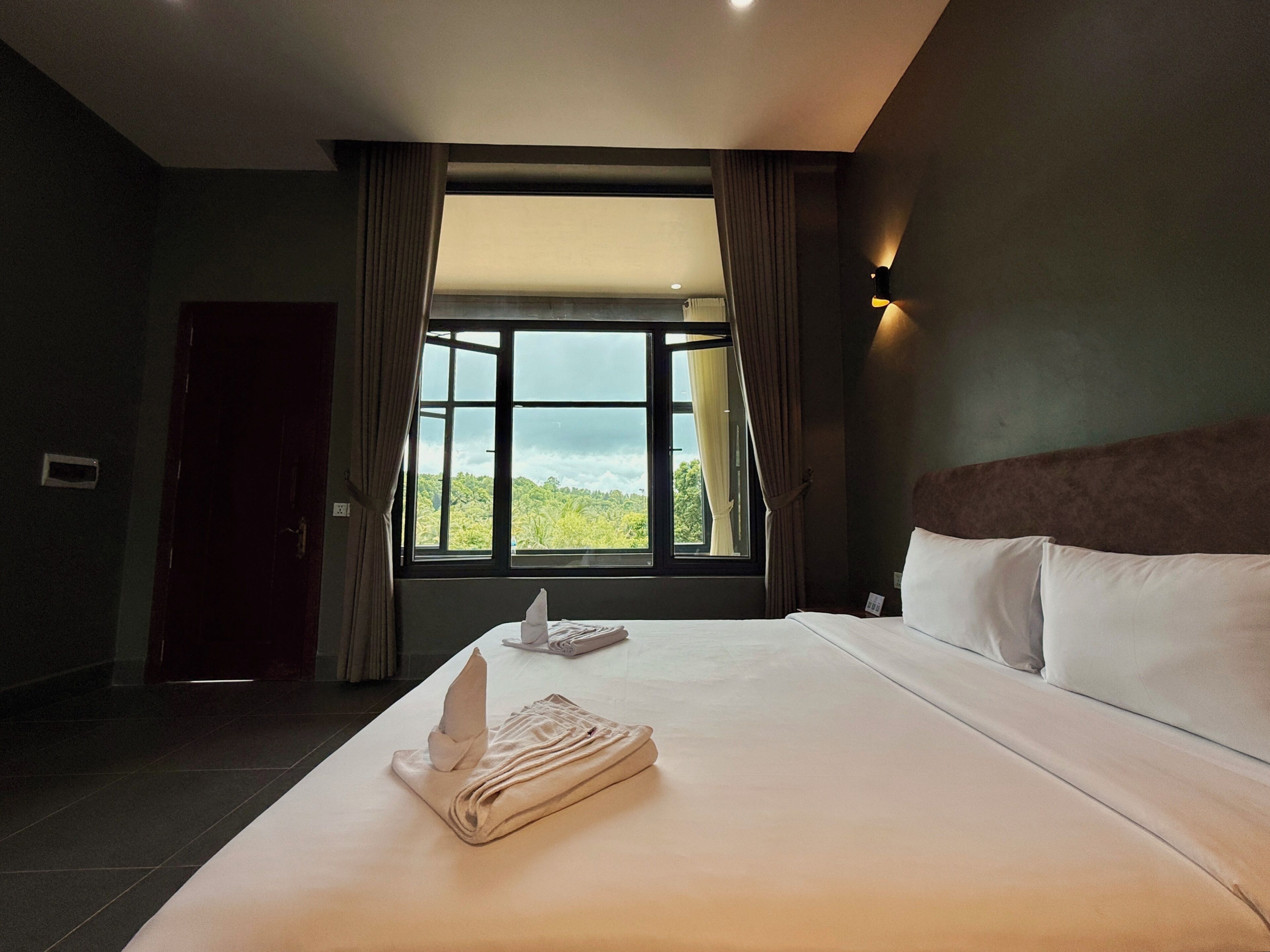A hotel room with a large bed, a window with curtains, a headboard, and a wall-mounted light fixture.