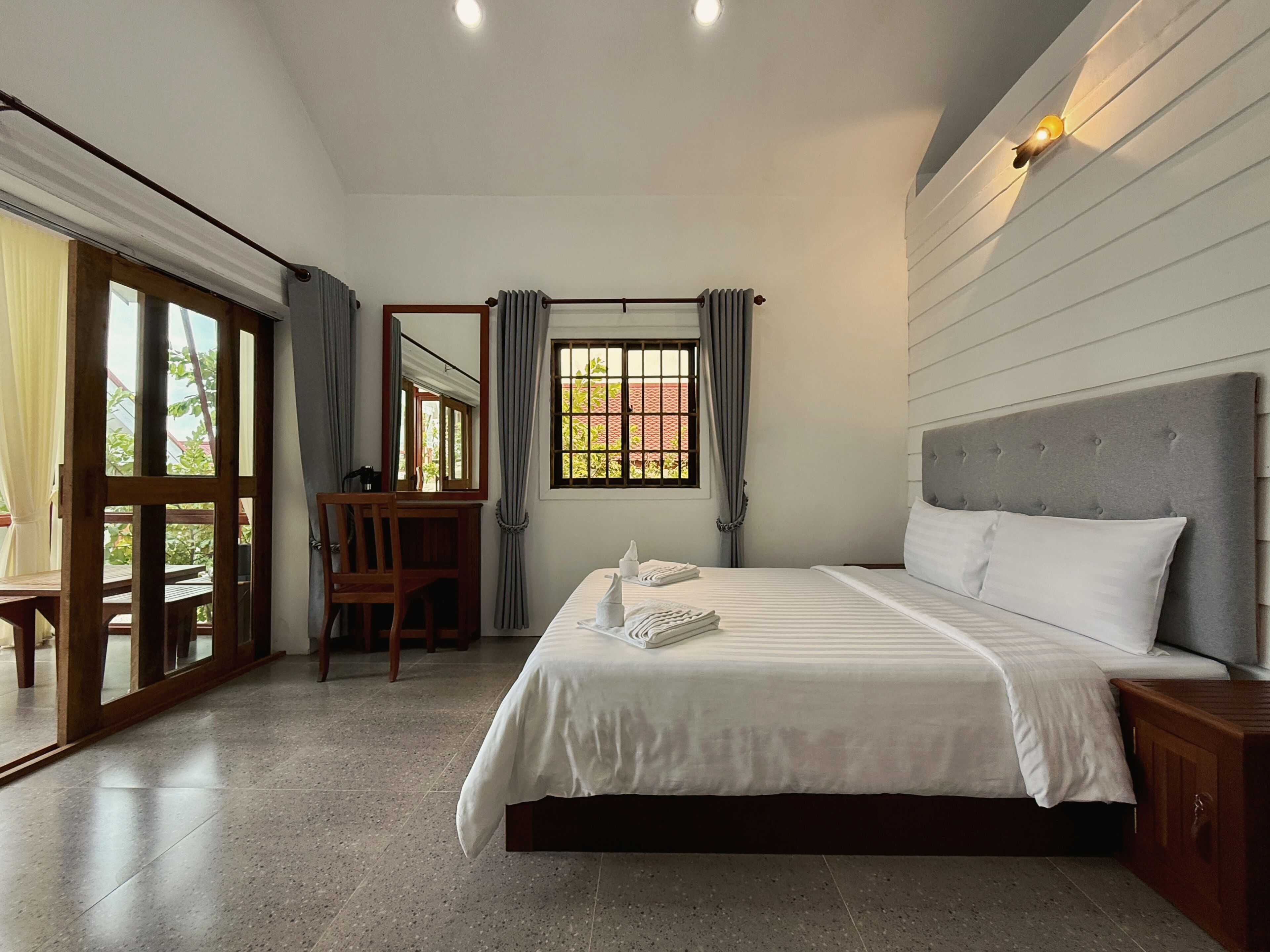 A bedroom with a bed, a desk, a chair, a window with curtains, and a view of greenery outside.