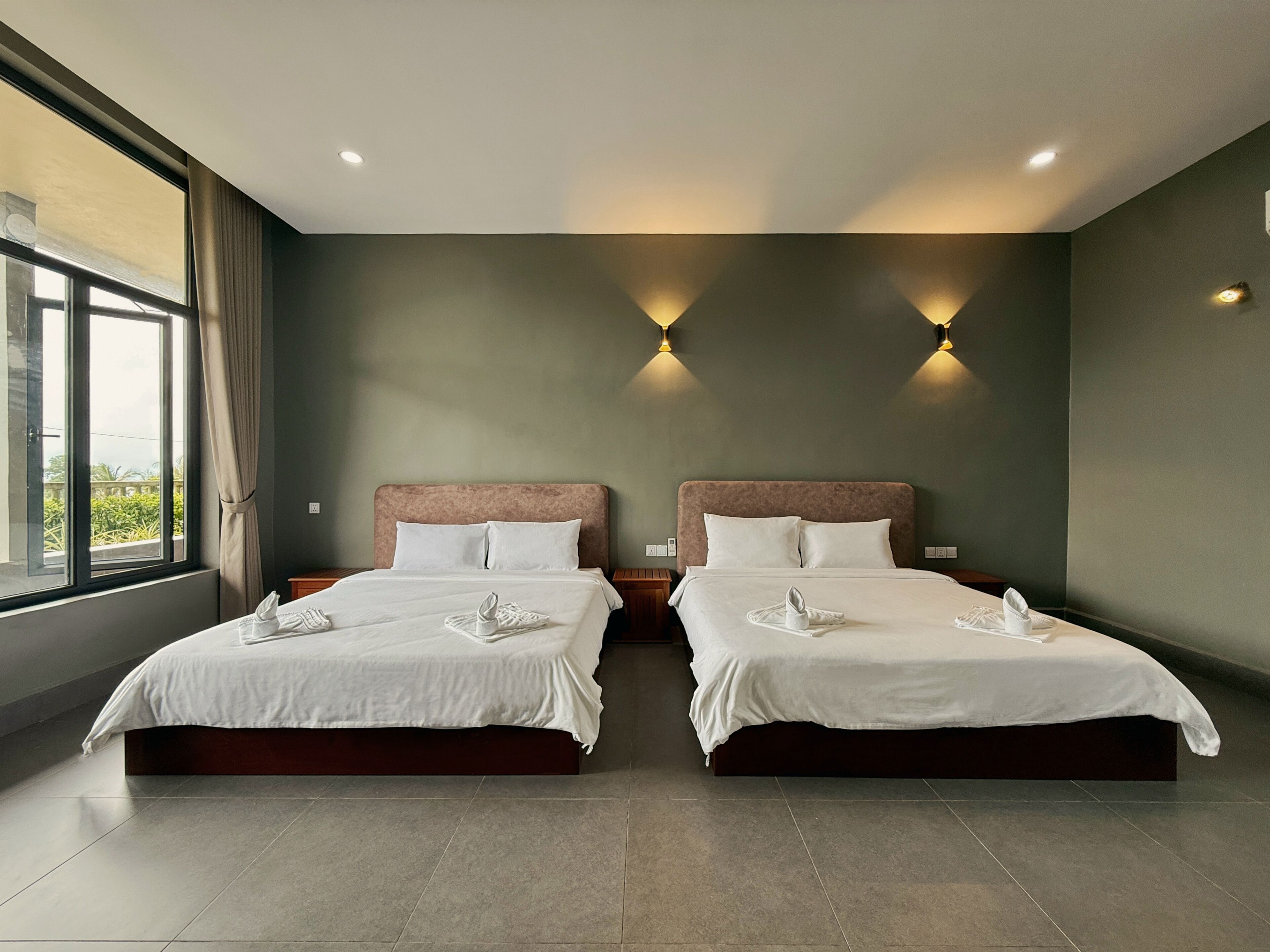 A hotel room with two beds, a large window, and wall-mounted lighting.
