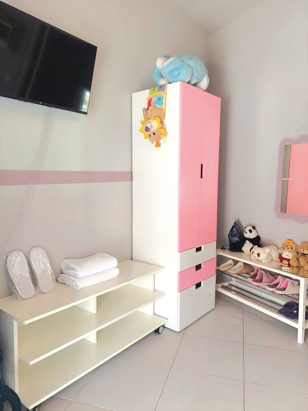 A child's bedroom with a pink and white wardrobe, a TV mounted on the wall, a shelf with slippers, and a shelf with stuffed toys.