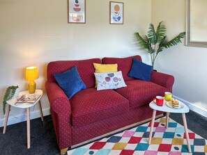 A red sofa with patterned cushions, a small side table with a lamp, a colorful rug, and a plant. - 2 bedroom accommodation in Perranporth (Perranporth)