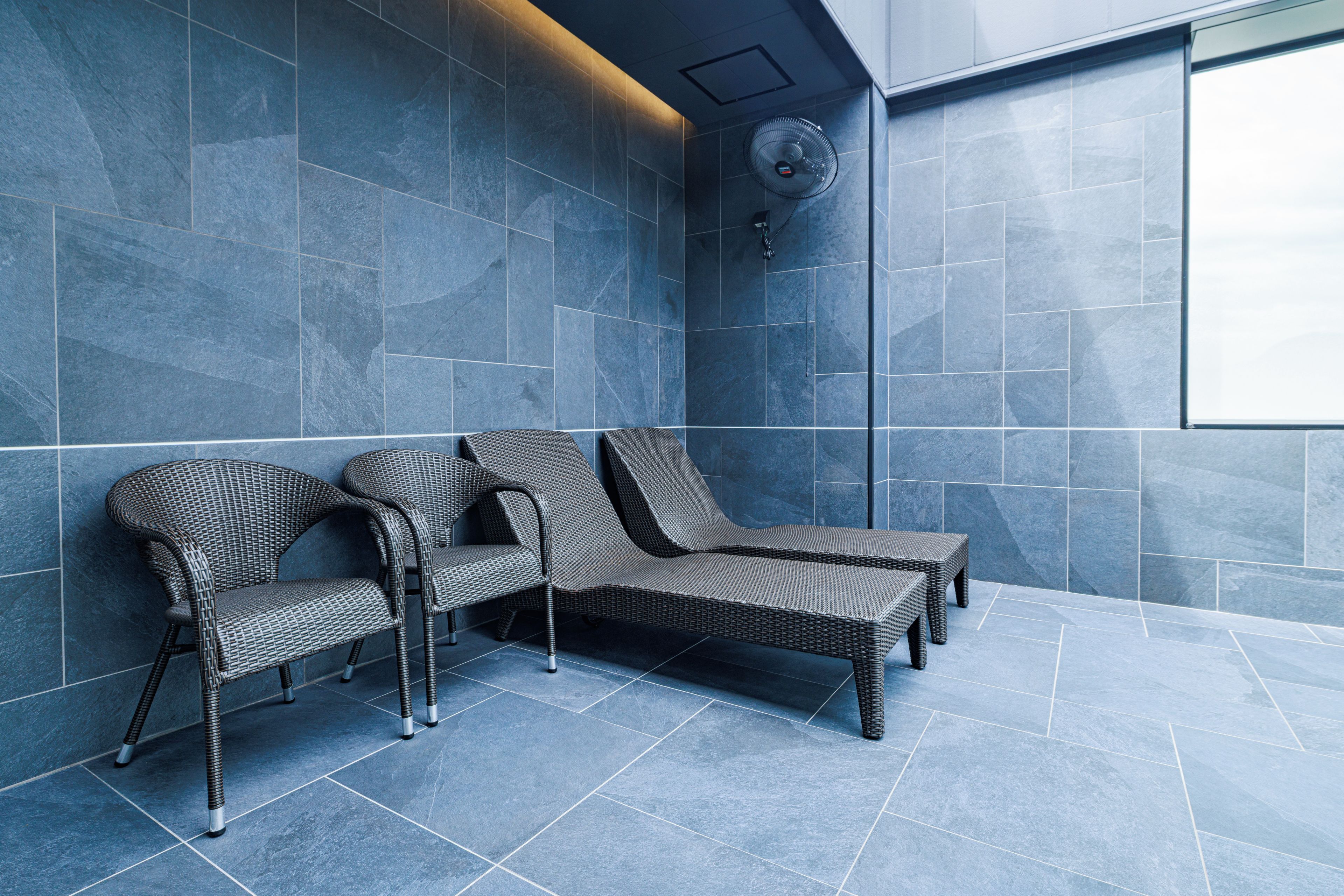 A modern spa area with dark blue tiled walls, two wicker lounge chairs, and a ceiling fan.
