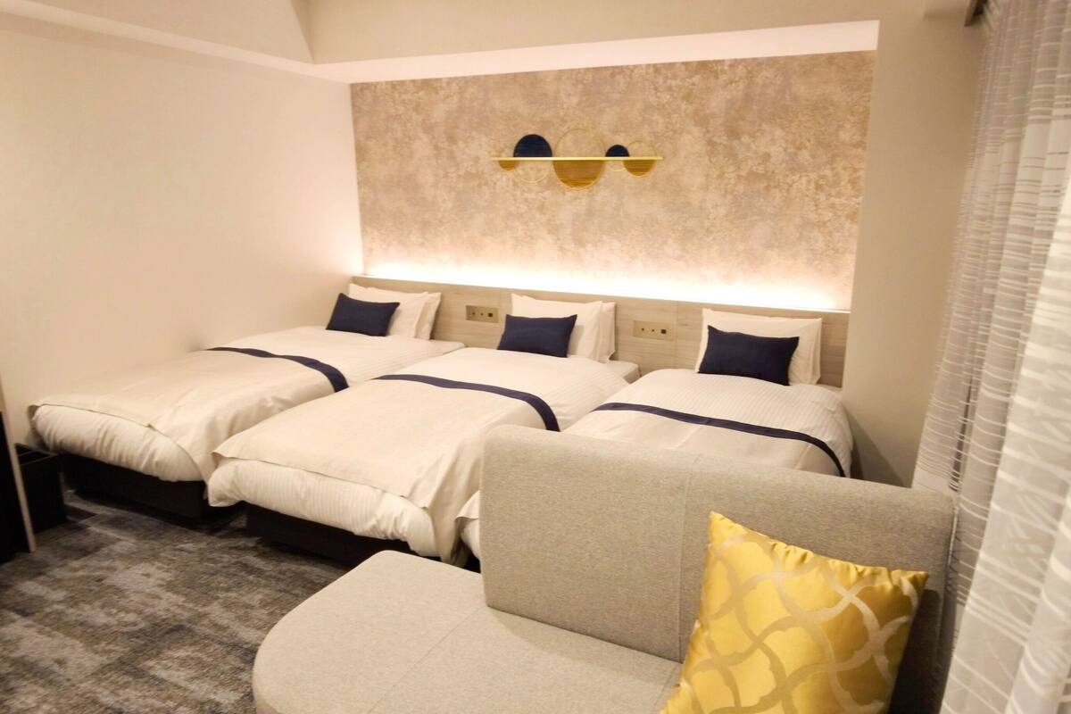 A hotel room with three beds, a sofa, a wall-mounted light fixture, and a window with blinds.