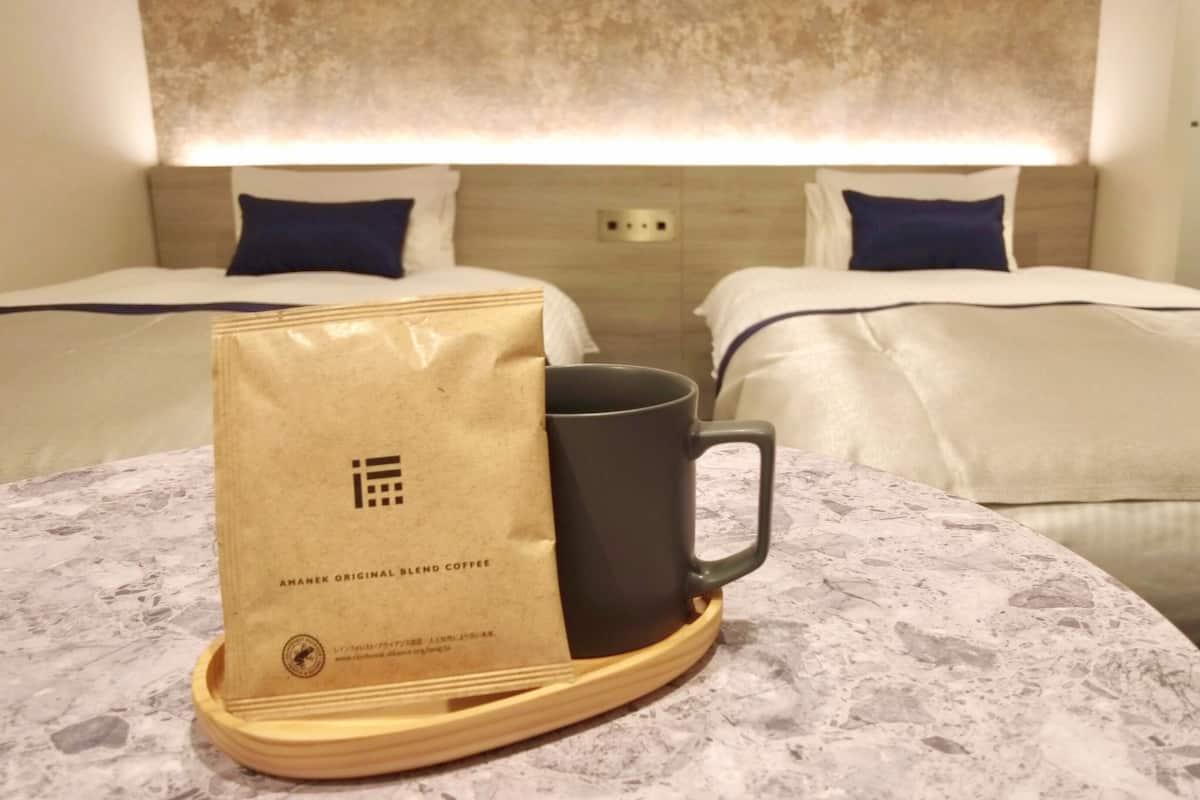 A hotel room with two beds, a table with a coffee bag and a mug, and a textured wall.