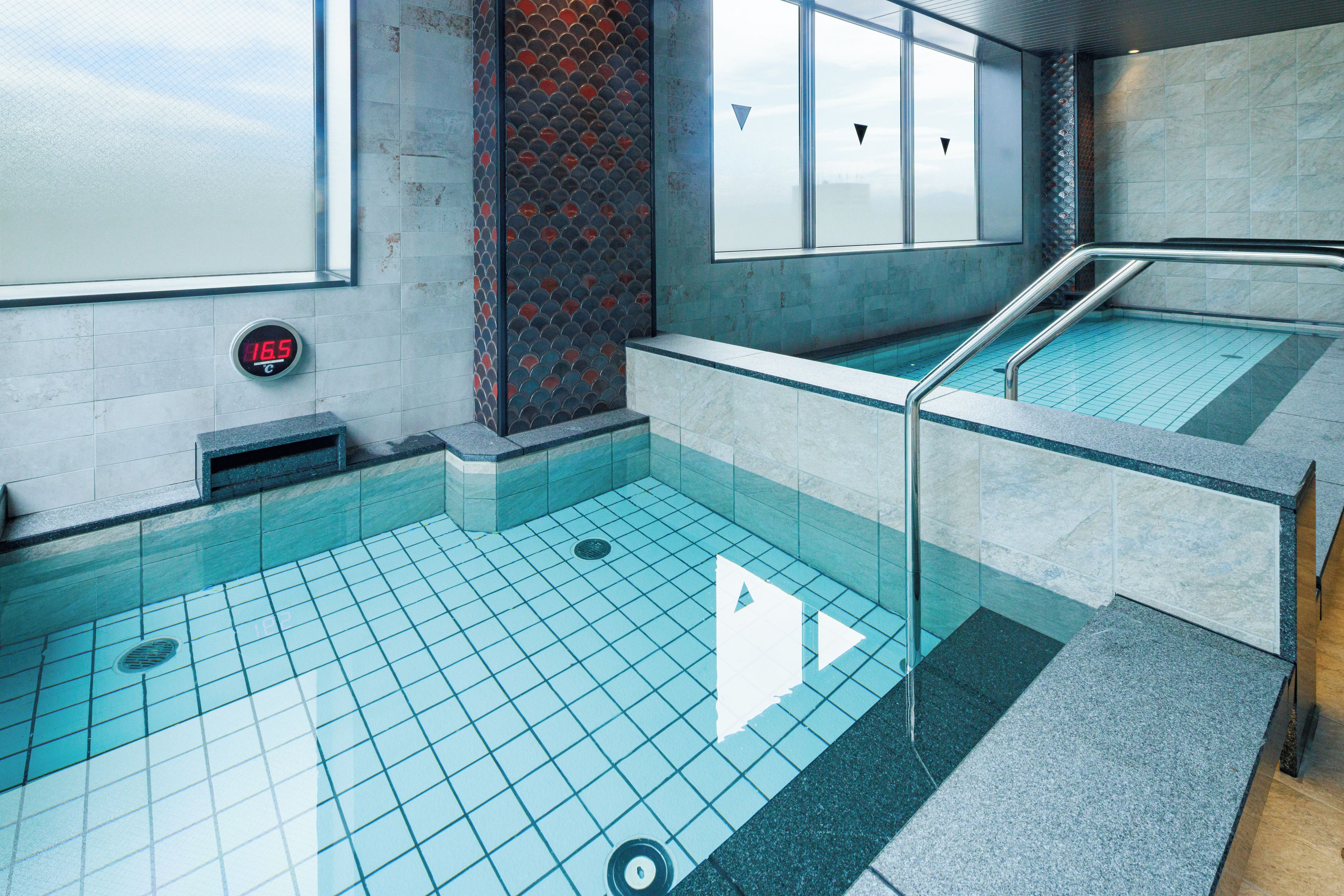 Uma piscina com piso ladrilhado, visor de temperatura e degraus que levam à água.