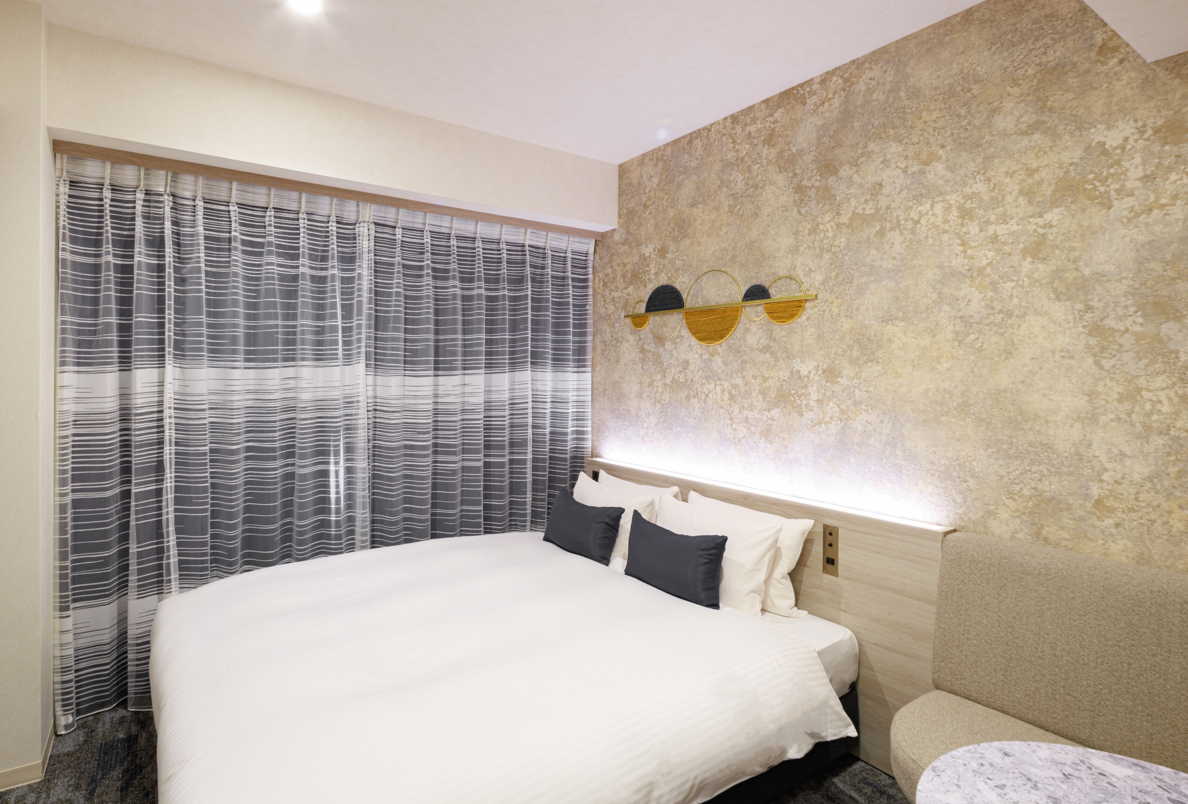 Ein modernes Hotelzimmer mit einem großen Bett, einem Kopfteil mit integrierter Beleuchtung, einer strukturierten Wand und einem Fenster mit gestreifteren Vorhängen.