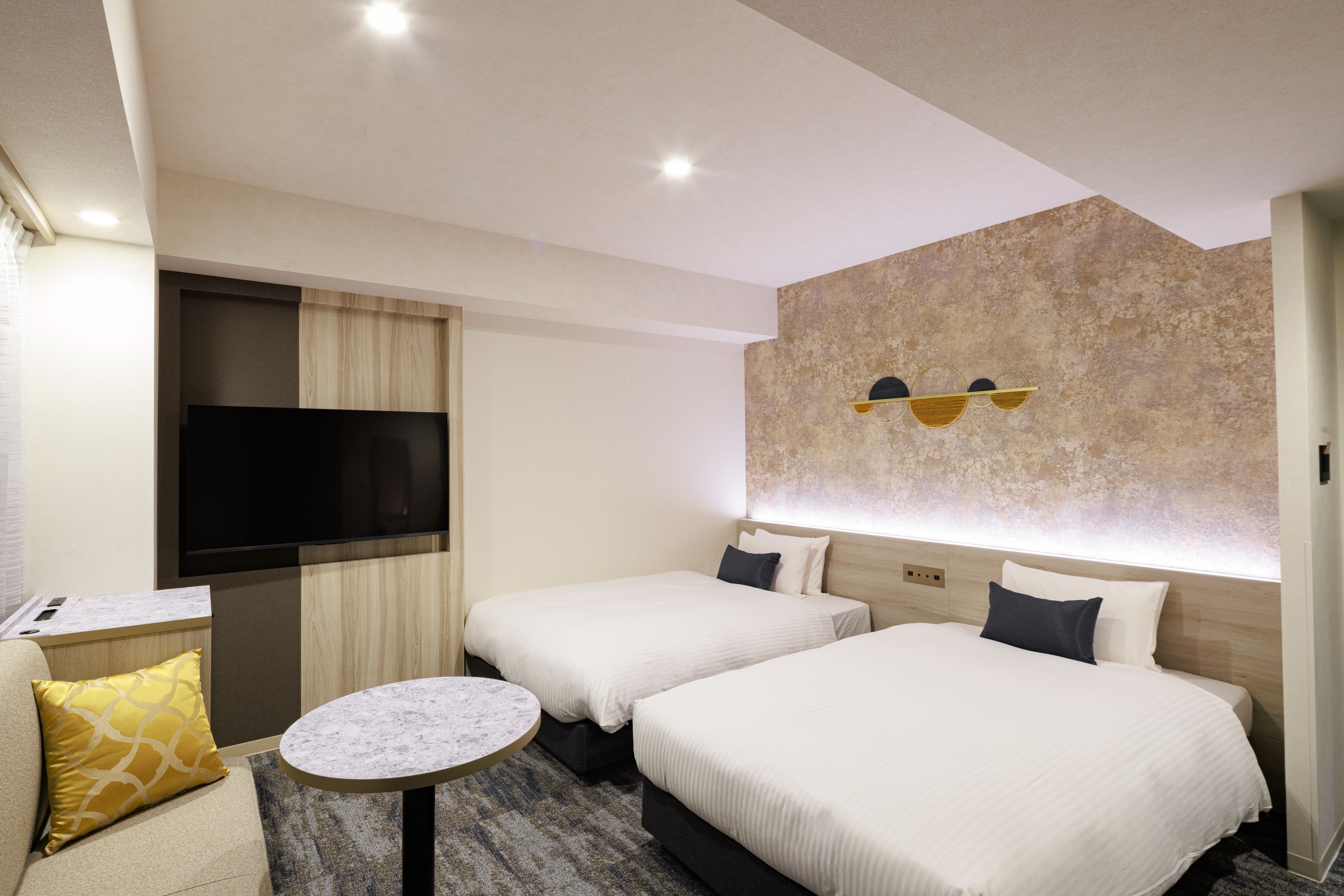 A hotel room with two beds, a TV, a small table, and a wall-mounted light fixture.