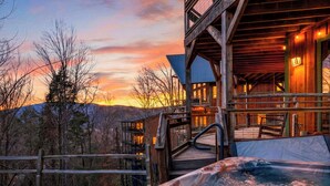 Een houten chalet met een jacuzzi en terras, omgeven door bomen en uitzicht op een zonsondergang.