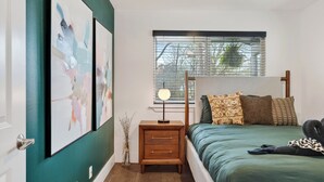 Quarto com cama, mesa de cabeceira, luminária, janela com persianas e obras de arte na parede.