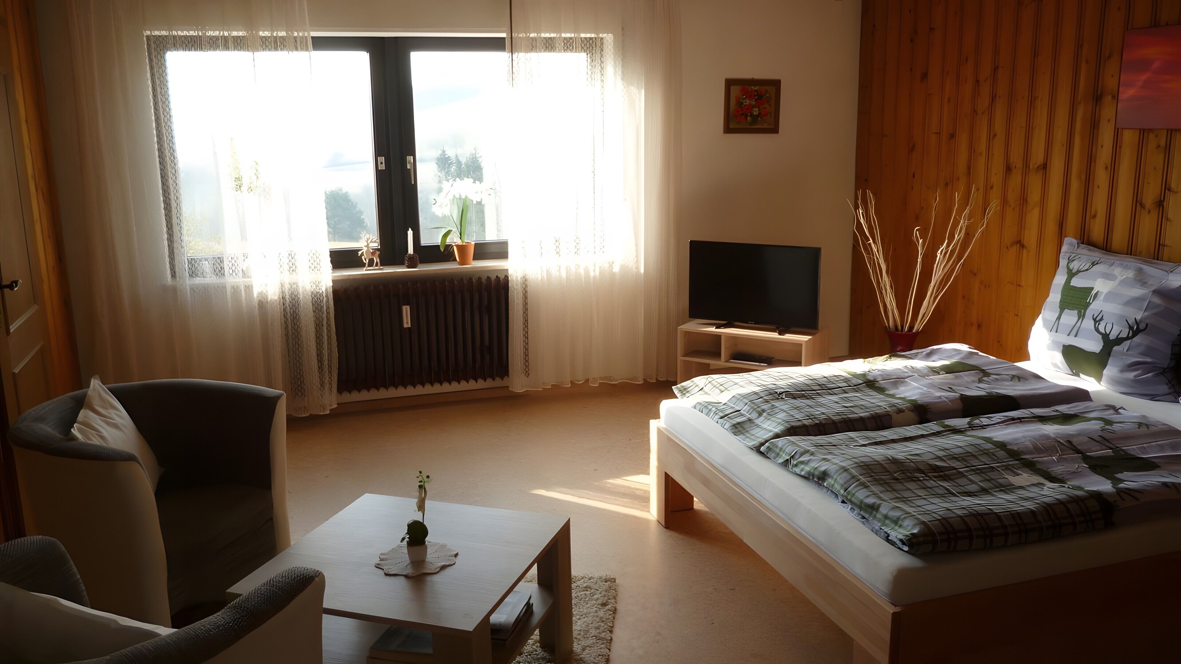 Ein Schlafzimmer mit einem Bett, einem Fernseher, einem Sessel, einem Tisch und einem Fenster mit Vorhängen.