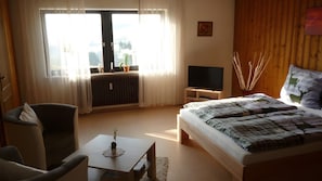 Ein Schlafzimmer mit einem Bett, einem Fernseher, einem Sessel, einem Tisch und einem Fenster mit Vorhängen.