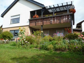 Ein zweigeschossiges Haus mit Balkon, umgeben von einem Garten mit verschiedenen Blumen und Pflanzen.
