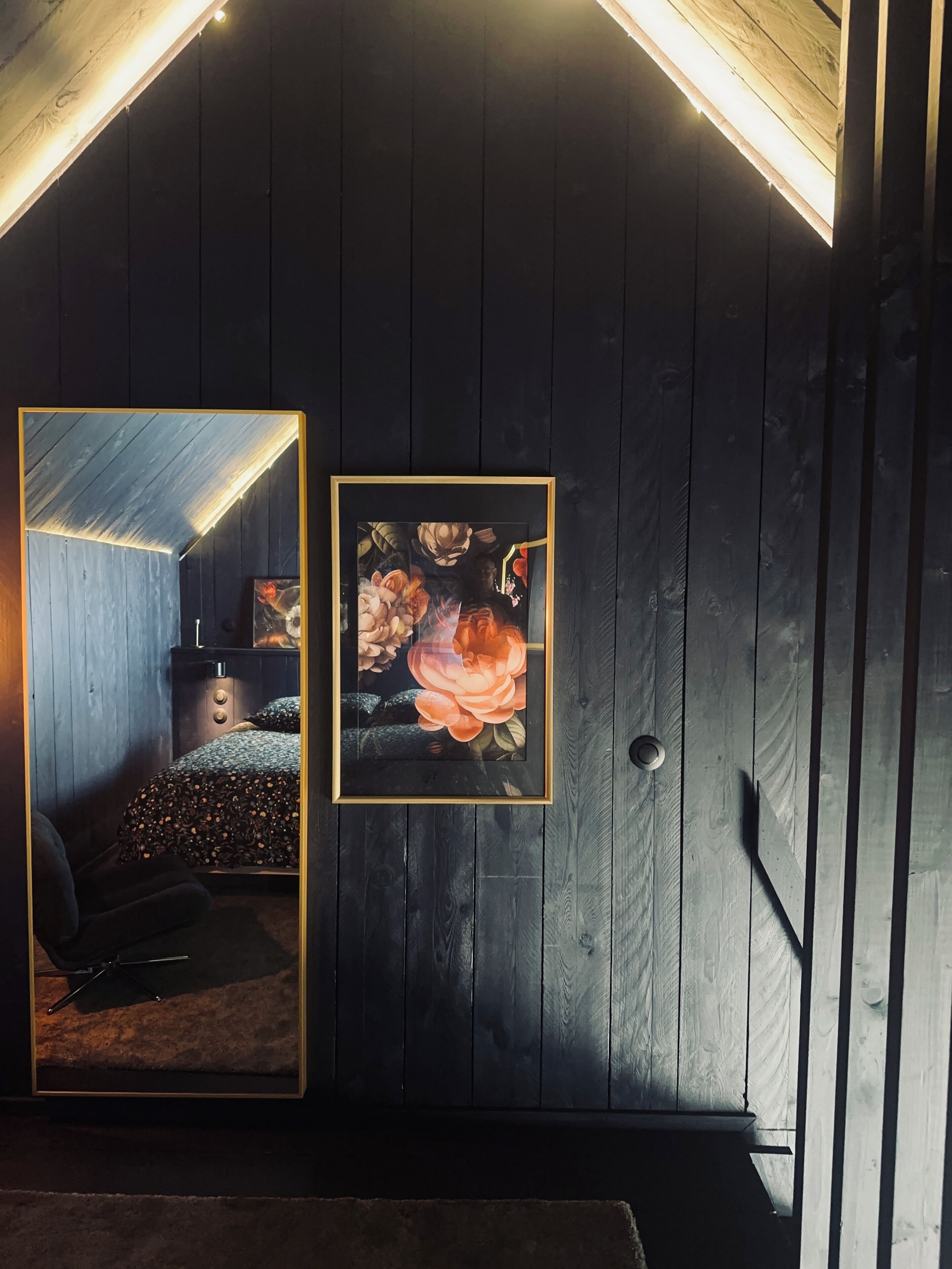 Une pièce sombre aux murs en bois, avec un plafond en pente, un lit recouvert d’une courtepointe à motifs et un tableau encadré accroché au mur.