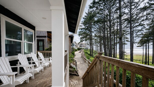 A wooden deck with white Adirondack chairs, a wooden railing, and a view of the ocean and trees.
