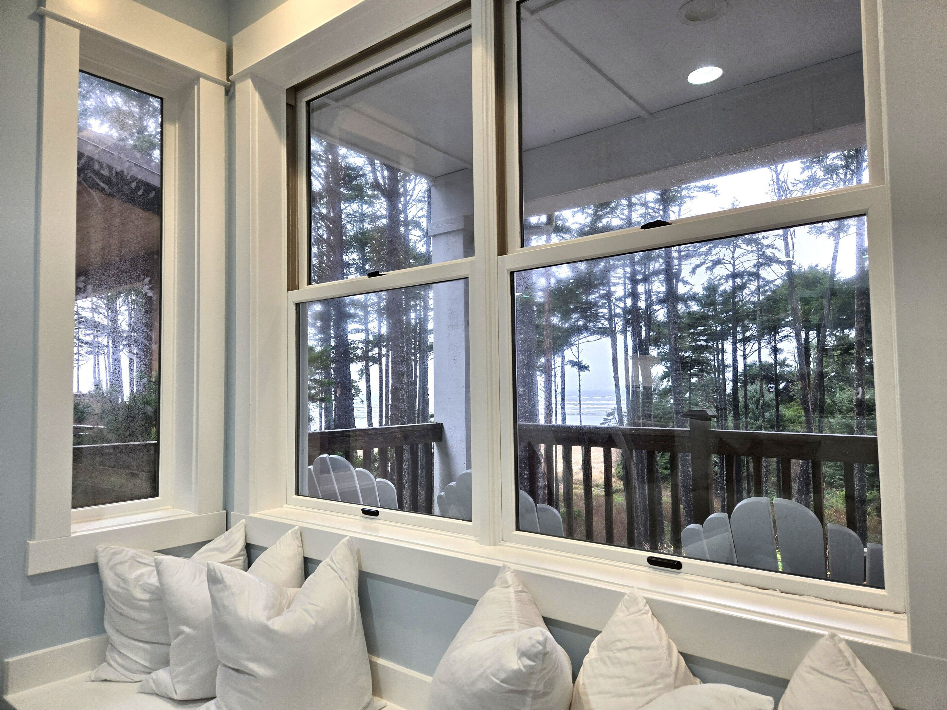 Una habitación con ventanas amplias, vistas a un bosque y una zona de estar con cojines blancos.
