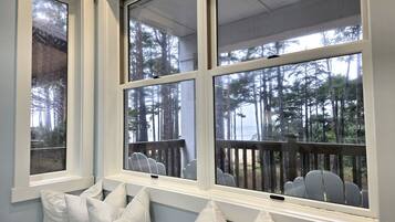 Una habitación con ventanas amplias, vistas a un bosque y una zona de estar con cojines blancos.