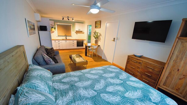 Une chambre à coucher avec un lit, une commode en bois, un téléviseur et, au fond, une petite cuisine.