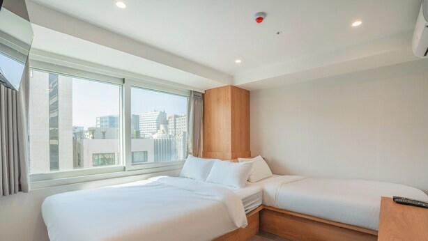 A modern hotel room with a large bed, a smaller bed, a wooden headboard, a desk, and a window with a city view. - KIMSTEL Jongno (Seoul)