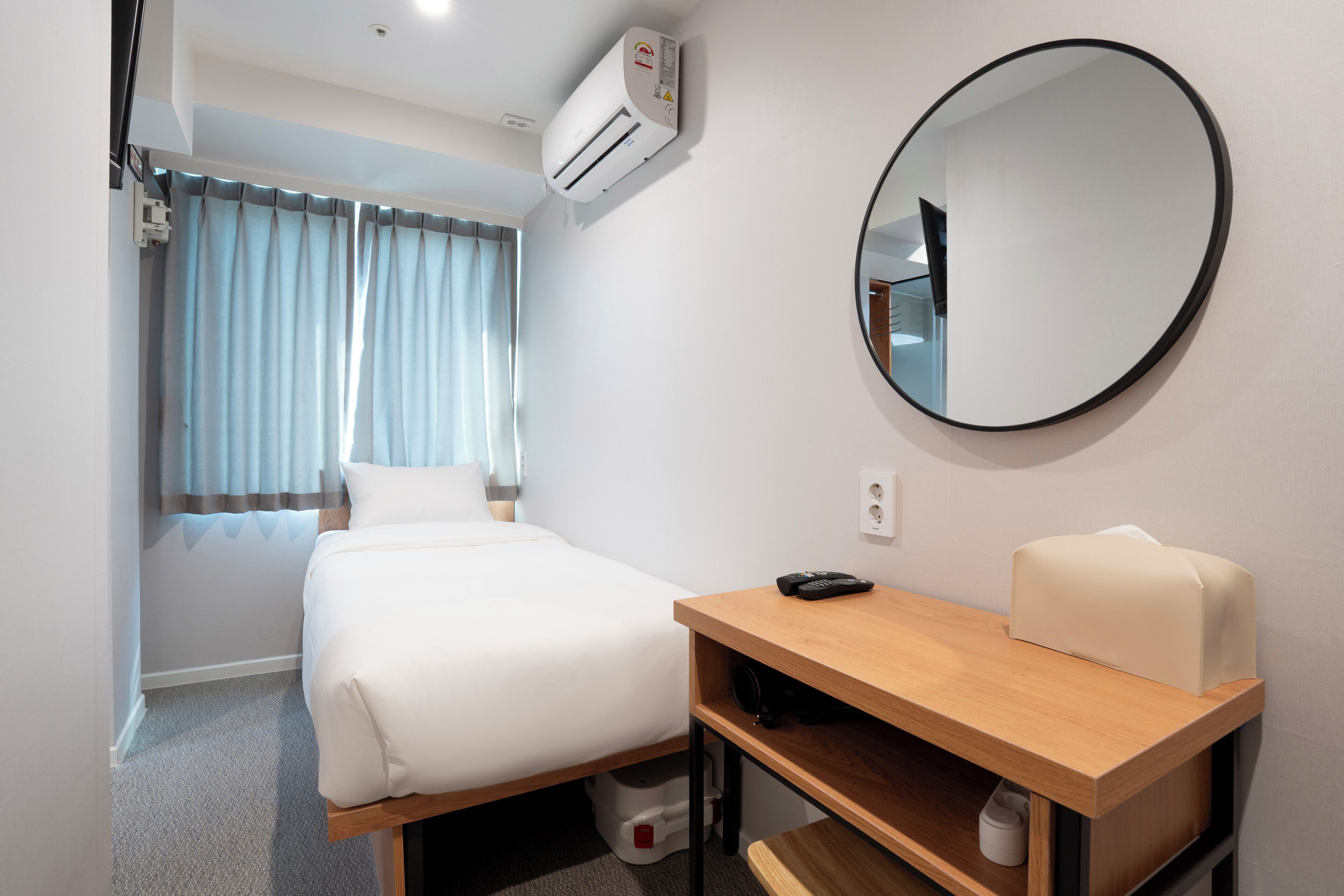 Une chambre d’hôtel comprenant un lit, un bureau avec une chaise, un miroir rond et un appareil de climatisation.
