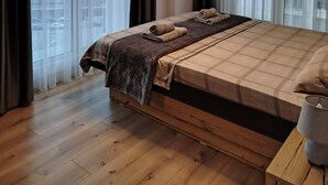 Een moderne slaapkamer met een groot bed, houten vloeren, een flatscreen televisie aan de muur en grote ramen met dunne gordijnen.