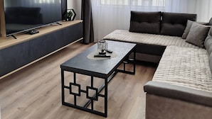 Een moderne woonkamer met een modulaire bank, een flatscreen televisie, een salontafel en een groot raam met gordijnen.
