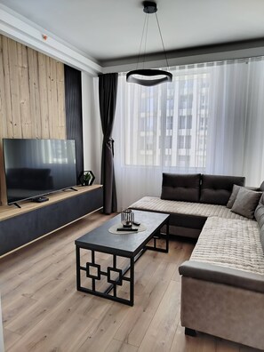 Ein modernes Wohnzimmer mit einem Ecksofa, einem Flachbildfernseher, einem Couchtisch und einem großen Fenster mit Vorhängen.