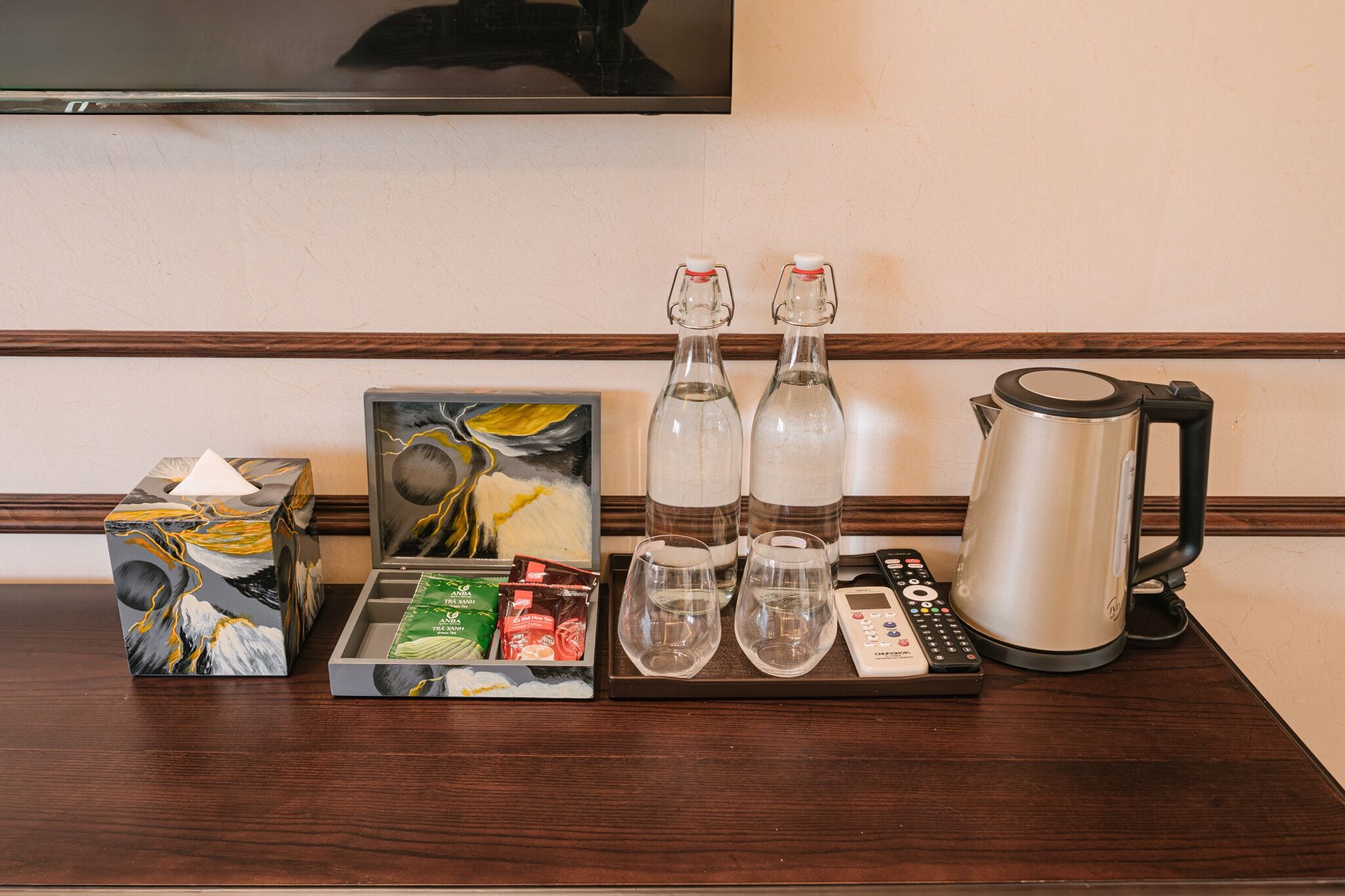 Hotellihuoneen minibaari, jossa on paperipyyhepakkaus, taulu, kaksi pulloa, kaksi lasia, kaukosäädin ja vedenkeitin.