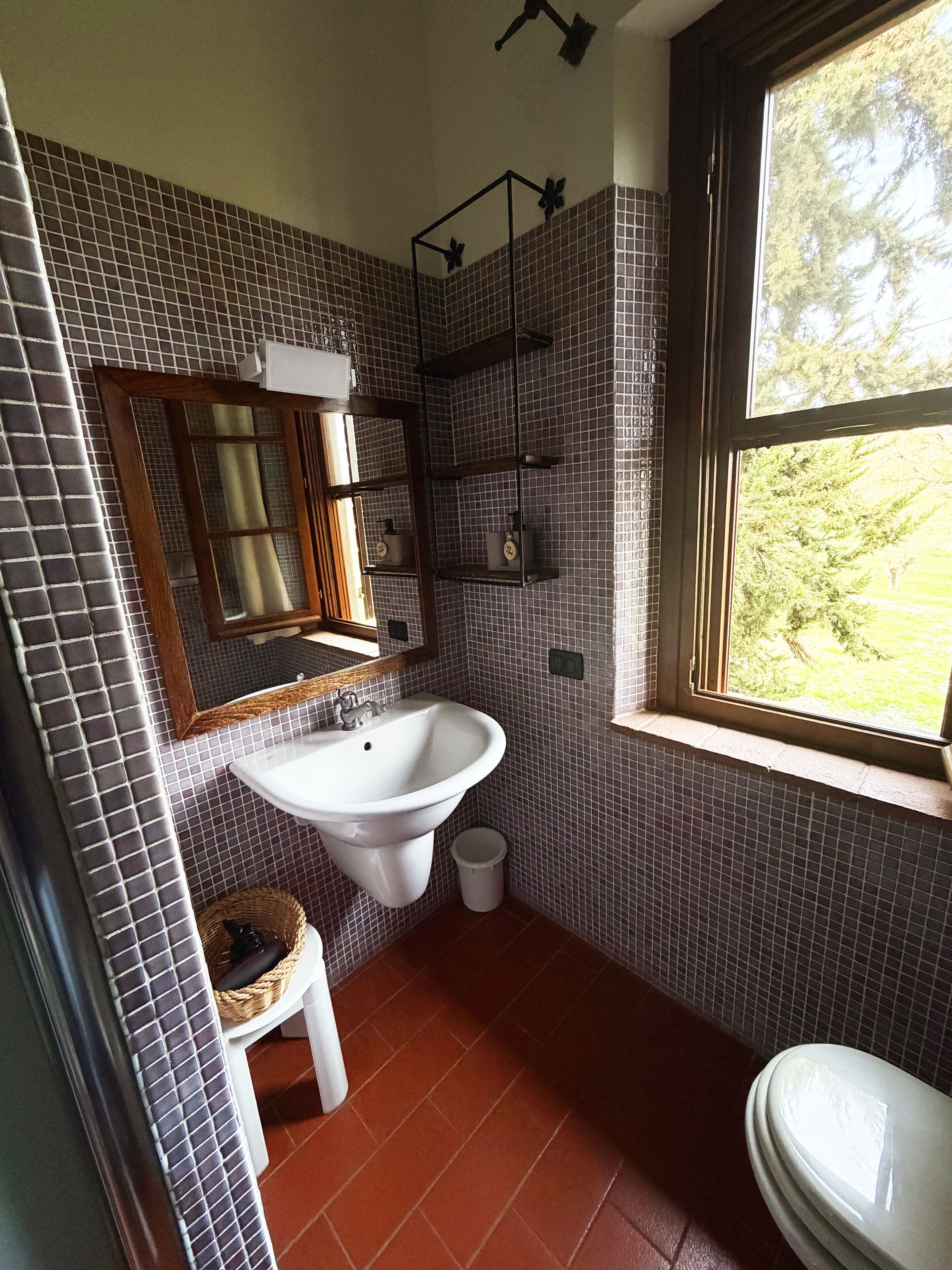 Um banheiro com parede revestida de azulejos, pia, espelho, janela e vaso sanitário.