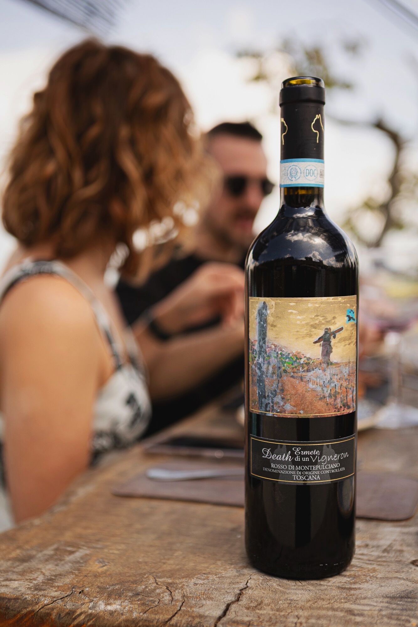 Una botella de vino con etiqueta que presenta una pintura de una figura en una colina, colocada sobre una mesa de madera.