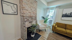 Ein Wohnzimmer mit Kamin aus Backstein, einem weißen Sessel, einem gelben Sofa und einem großen Fenster mit Vorhängen.