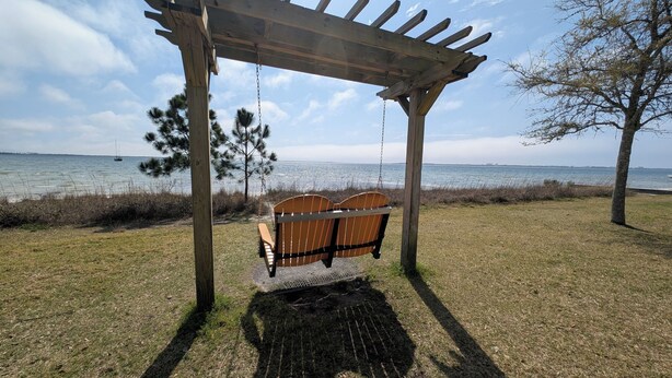 A wooden swing set with a metal seat and backrest, positioned under a wooden pergola, overlooking a body of water with a distant shoreline. - Miss Corby's Porch Swing | Historic St. Andrews (Panama City)