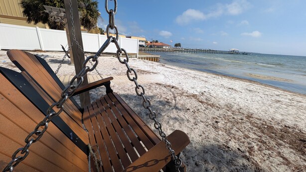 Eine Holzbank mit Metallketten, ein sandiger Strand und ein Steg, der ins Wasser hineinreicht.