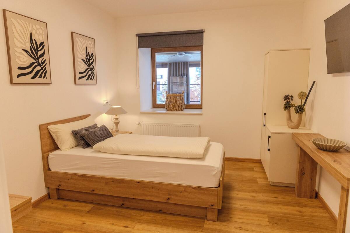 Une chambre avec un lit en bois, une lampe de chevet, une fenêtre donnant sur un beau paysage et une petite table sur laquelle est posé un vase.