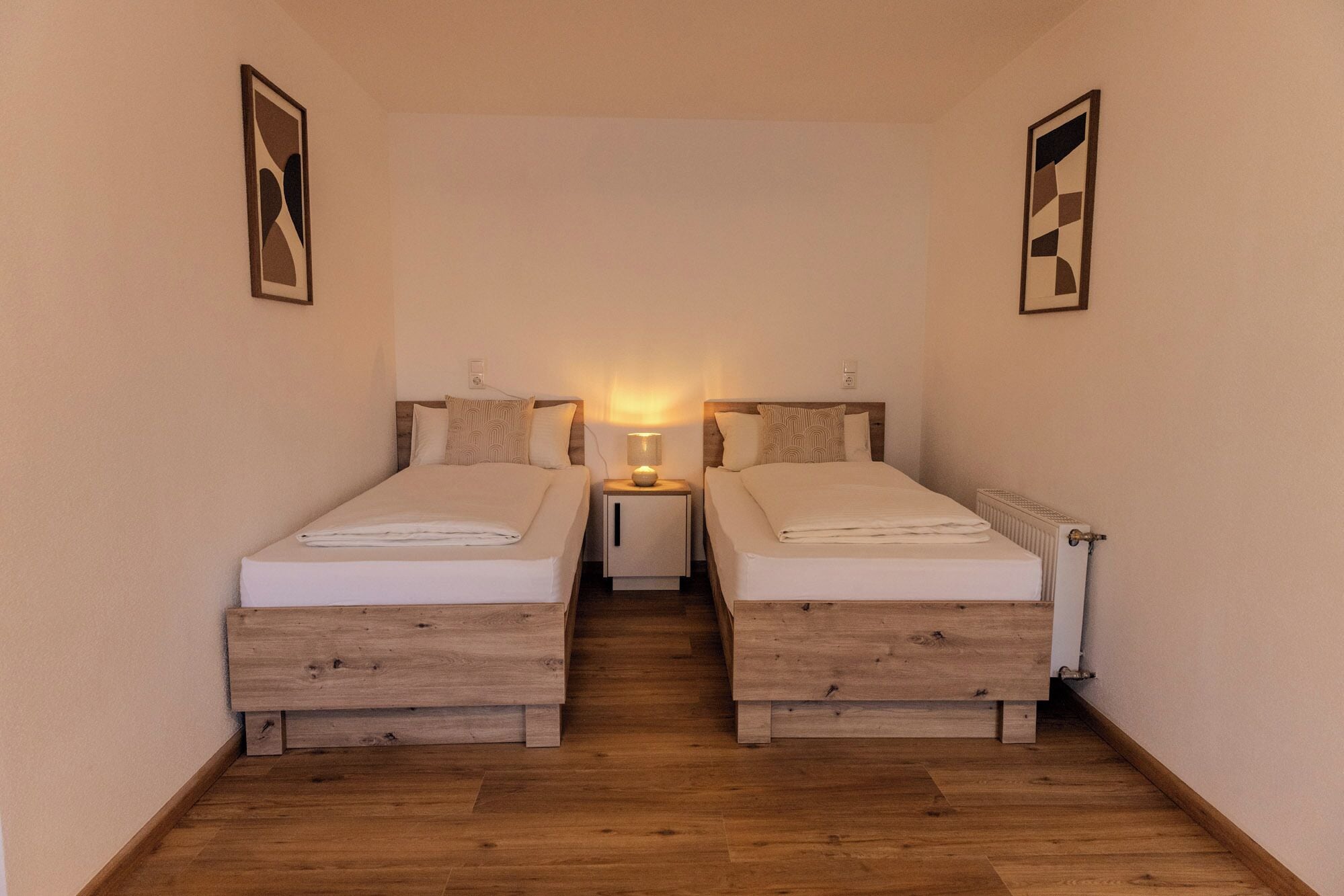 A narrow room with two beds, a nightstand with a lamp, and a radiator.