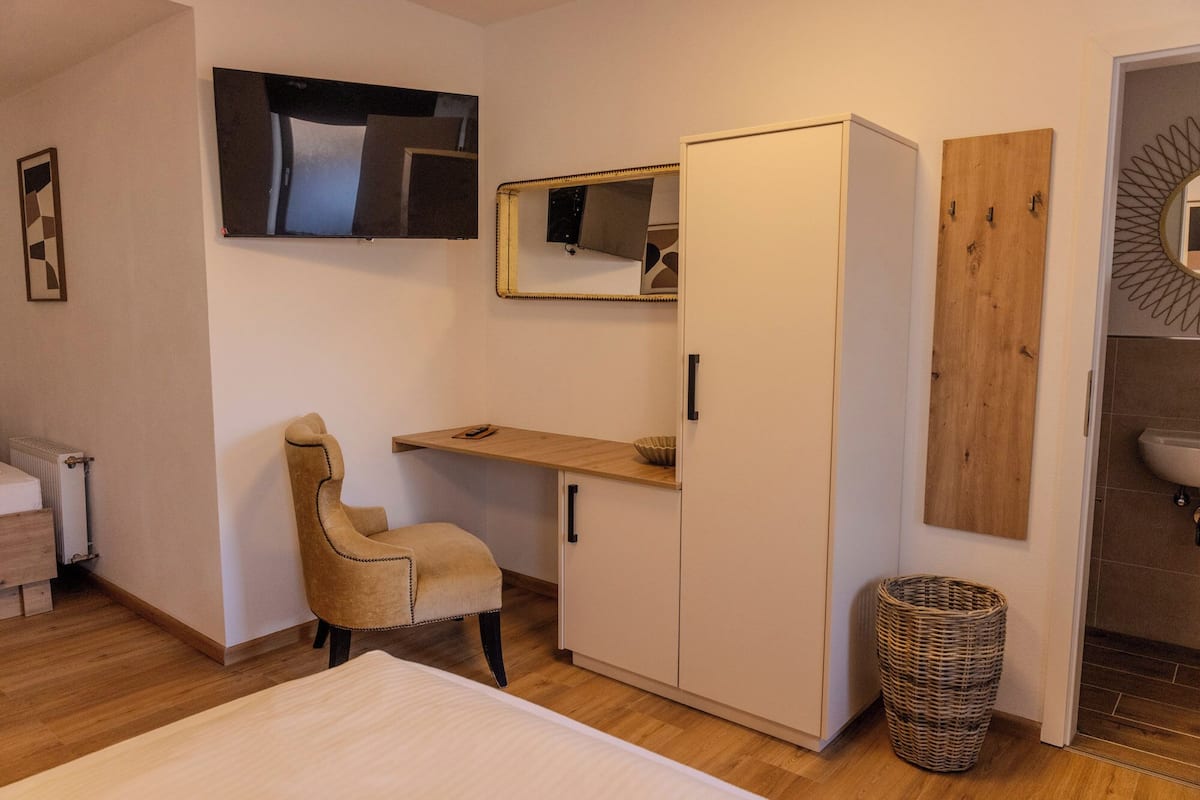 Une chambre à coucher comprenant un lit, une armoire, un bureau, une chaise, un miroir et un radiateur, ainsi qu’une salle de bain avec un lavabo et un miroir.