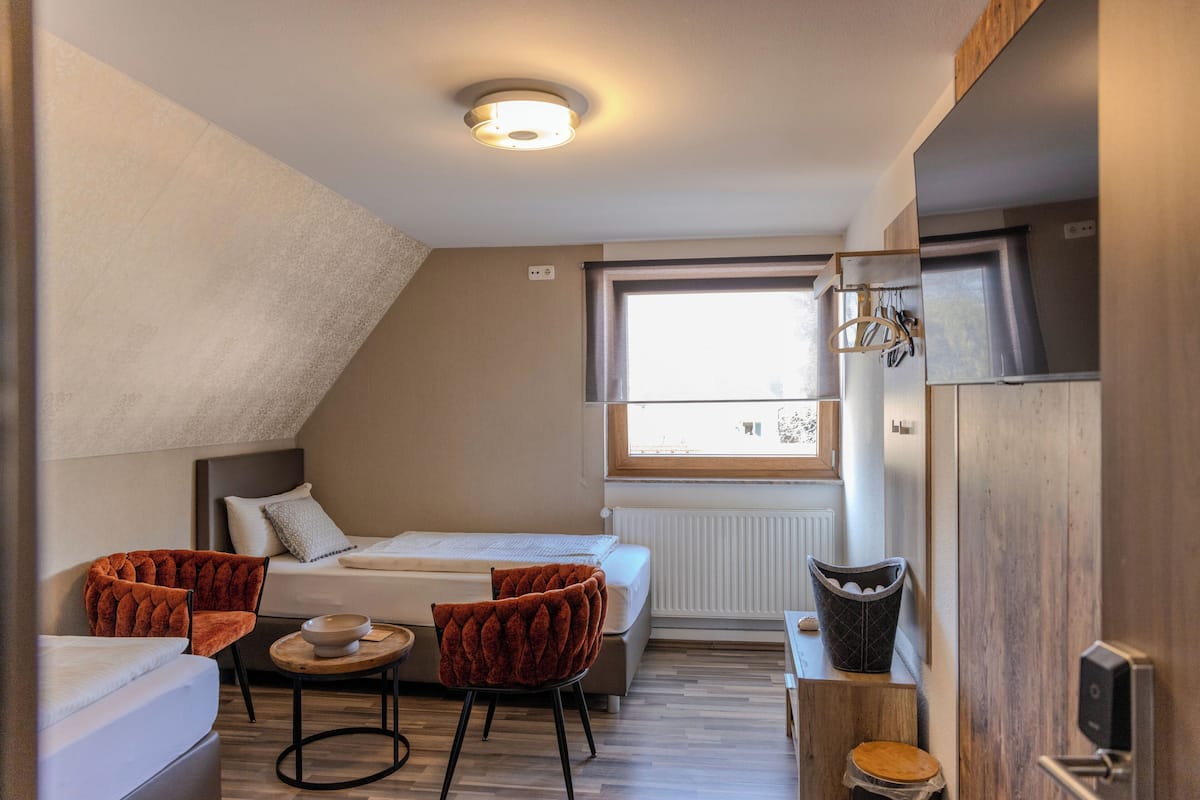Une petite pièce comprenant un lit, deux chaises, une petite table, une fenêtre et un radiateur.