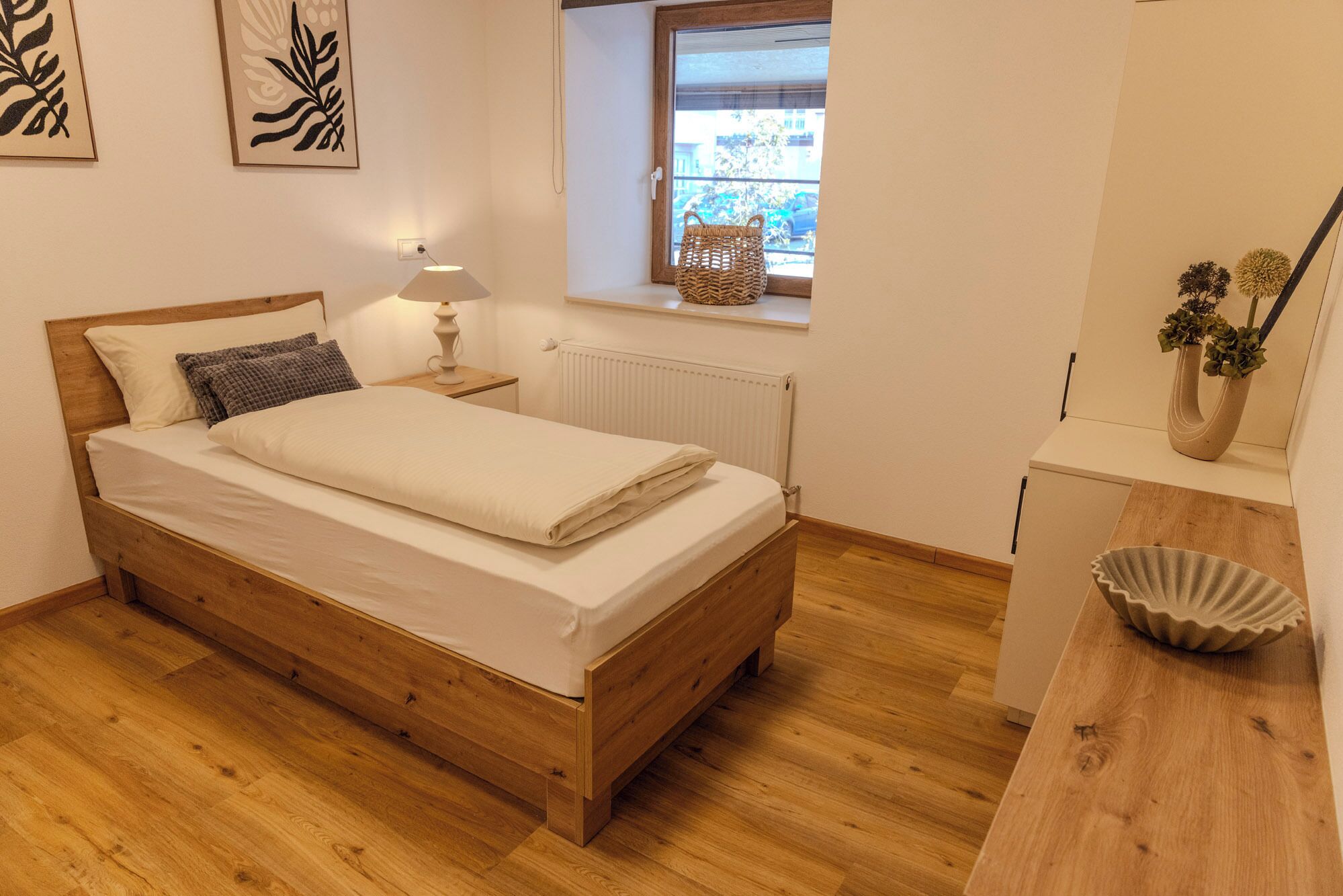 A bedroom with a wooden bed, a nightstand with a lamp, a radiator, a window with a view, and a small cabinet.