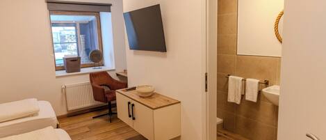 Habitación de hotel con piso de madera, cama con ropa blanca, pequeño escritorio con un tazón, una silla, ventana con vistas, televisor de pantalla plana y baño con lavamanos y perchero para toallas.